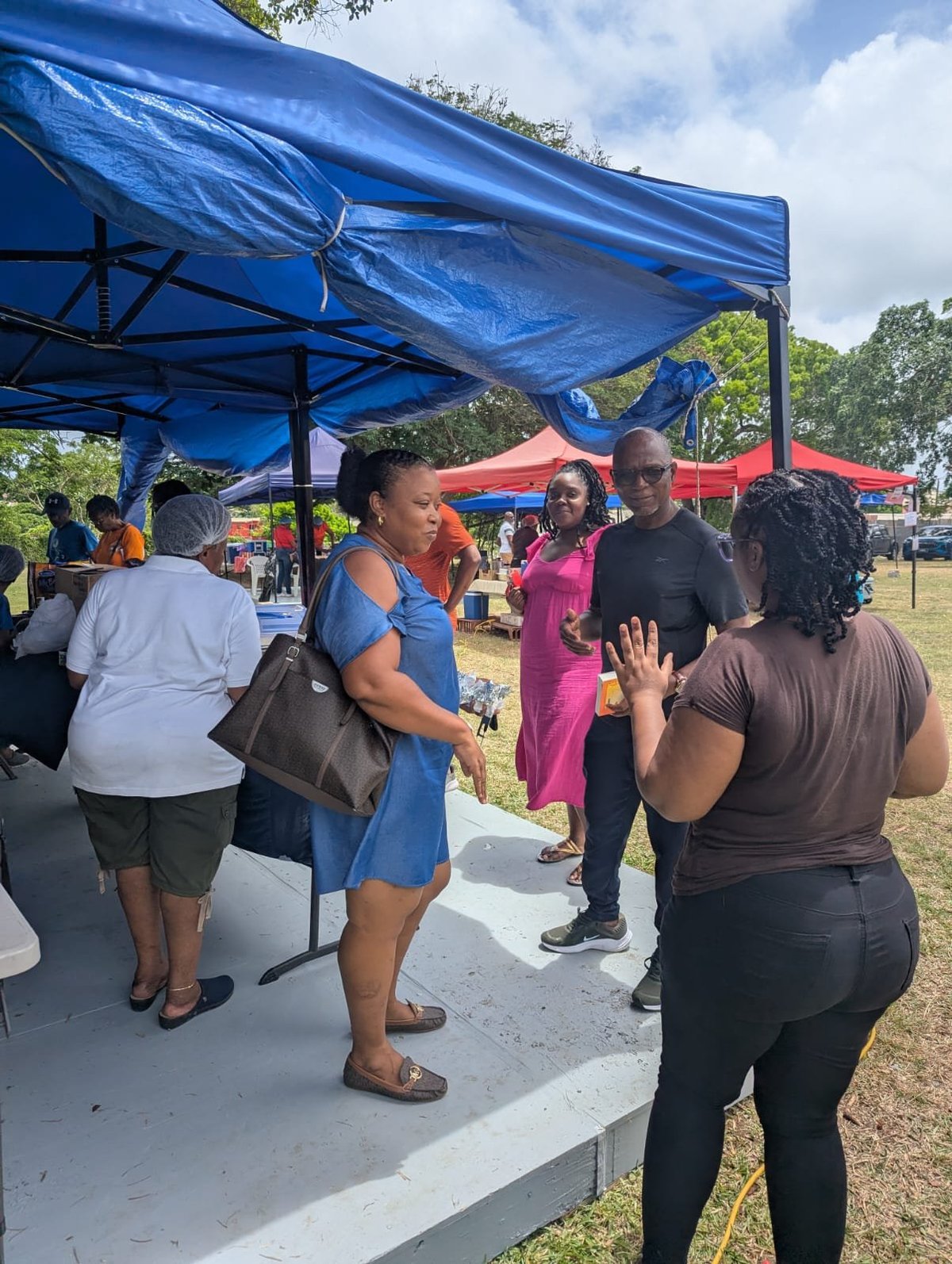 Group of people gathered under blue and red pop-up tents at the outdoor community event in a grassy field