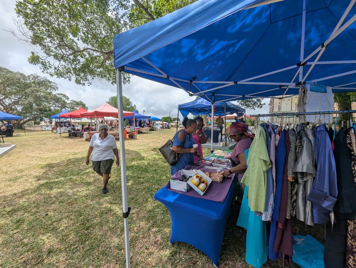 Outdoor market with vendors under a blue tent canopy selling clothing and goods on a green field, with multiple market stalls in the background