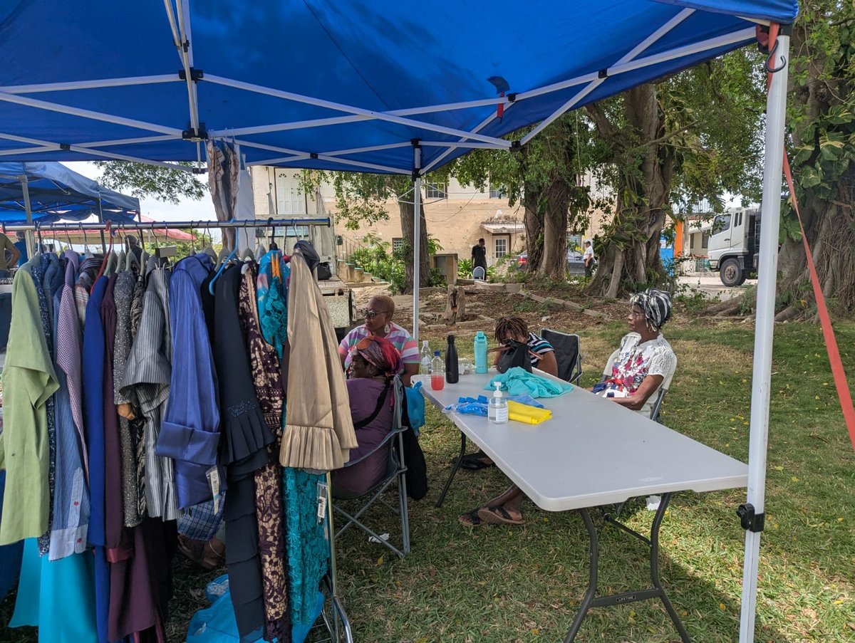Outdoor yard sale under a blue canopy tent with clothing racks on one side and shoppers browsing merchandise at a table