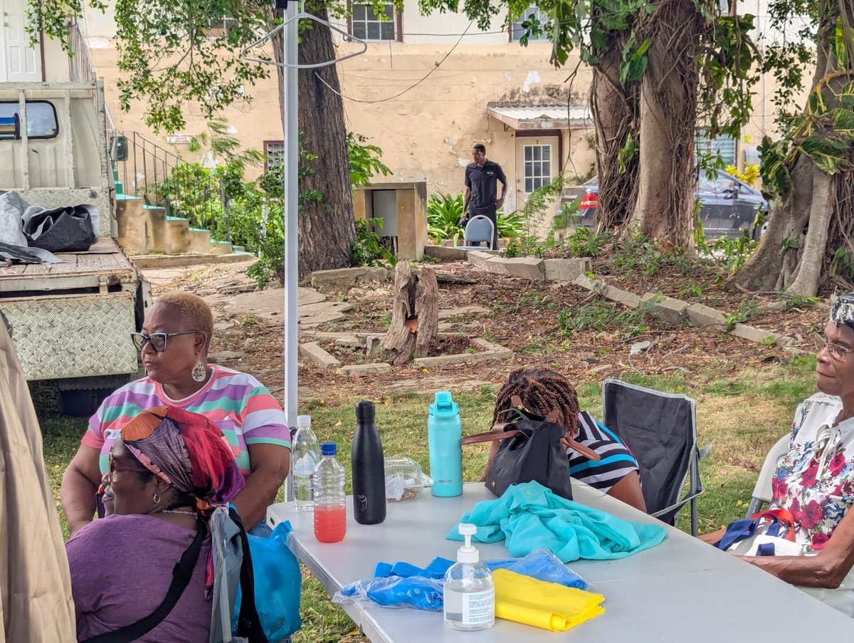 People gathered outdoors around a picnic table in a shaded backyard area with large trees and brick buildings in the background