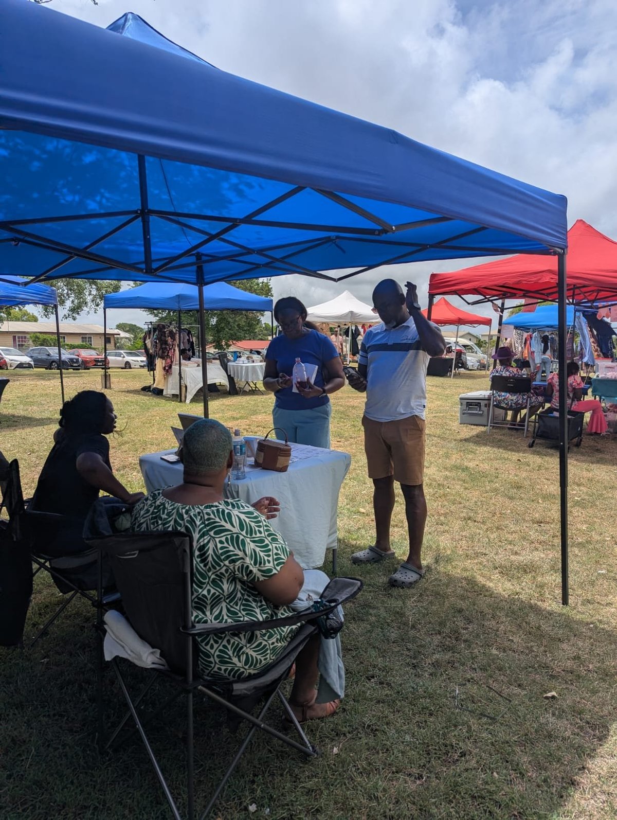 Outdoor community event under a blue tent canopy with people sitting and standing at booths on a grassy field
